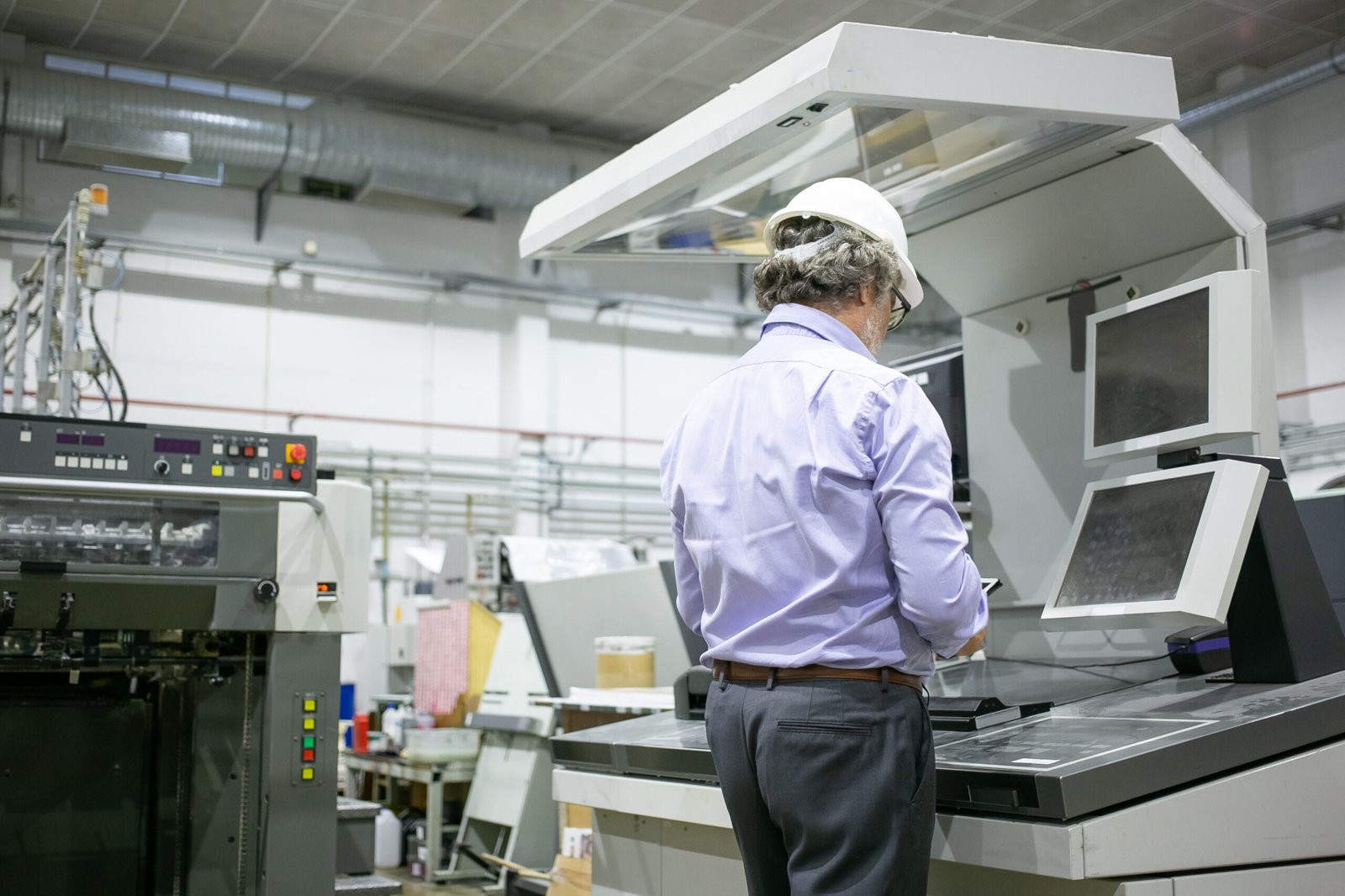 Grey haired male plant engineer in hardhat and glasses standing at industrial machine, using digital device. Copy space, back view. Industrial technology or machinery concept